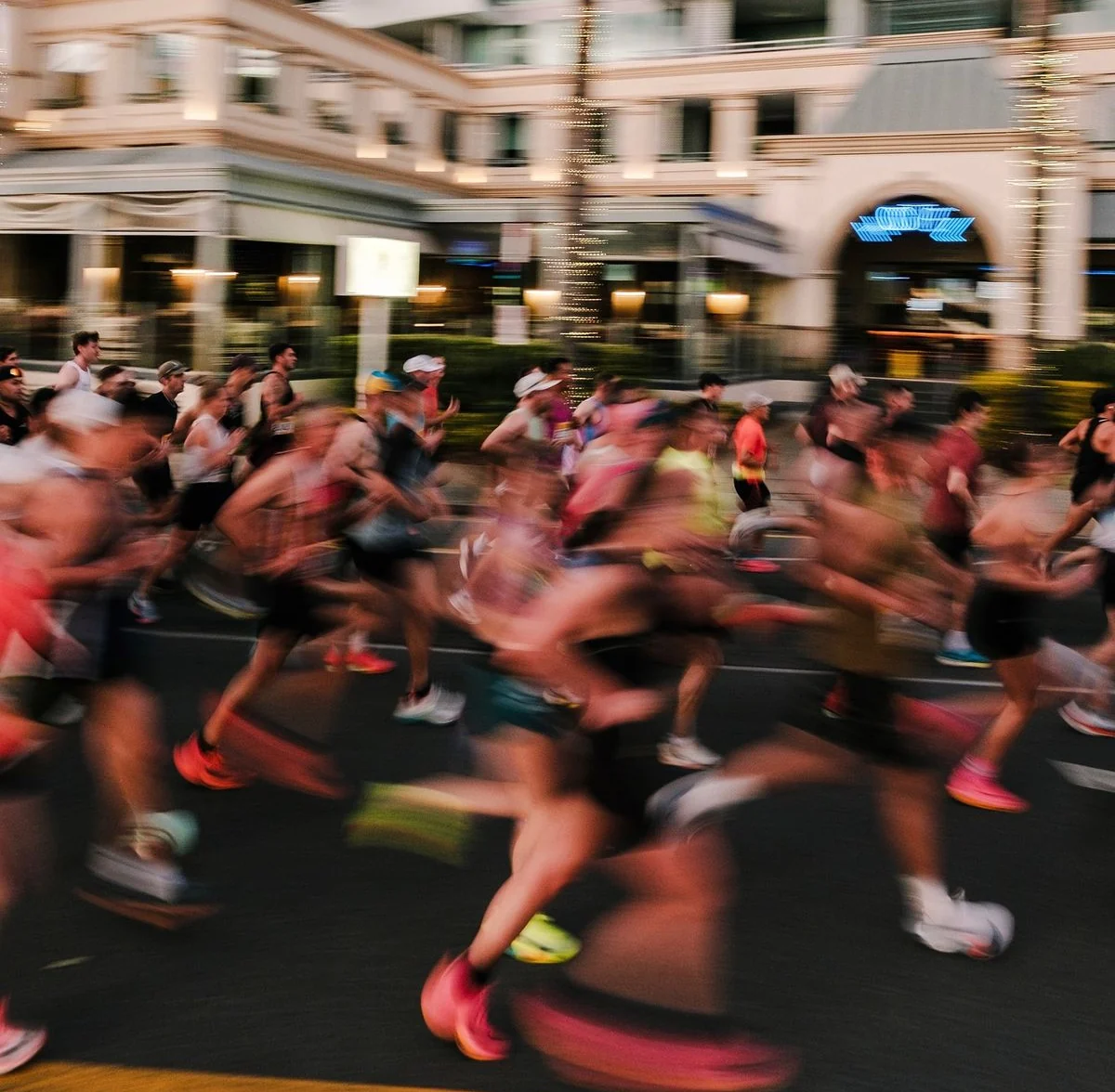 Pelotón de corredores en plena salida de una maratón urbana, en movimiento sobre la calle al atardecer, con un edificio y palmeras de fondo