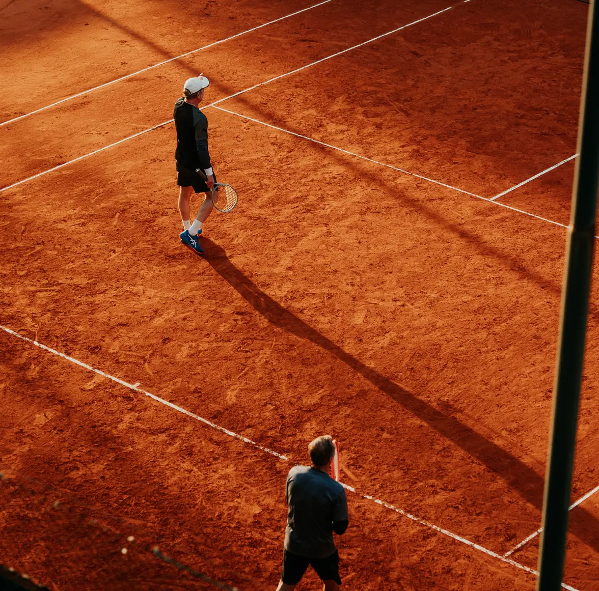 Vista desde lo alto de dos tenistas en una cancha de tierra batida al atardecer, con sus sombras largas sobre el polvo de ladrillo
