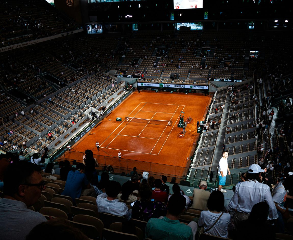 El Court Philippe-Chatrier en penumbra, con la tierra batida iluminada y el personal regando la cancha frente a las tribunas